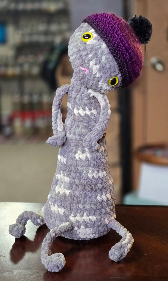 A crochet toy resembling a gray cat with a pink nose and bright yellow eyes, wearing a purple beanie hat.
